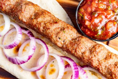 Lezzetli bir kebap, ince dilimler halinde kırmızı soğanla süslenmiş ve yanında, tadını çıkarmaya hazır, canlı bir salsa sosuyla sıcak ve yassı bir ekmeğin üzerinde oturur..
