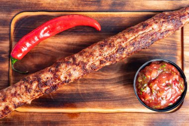 Sulu ızgara ızgara et ahşap bir tahtanın üzerinde dinleniyor. Yanında bir kase canlı salsa ve taze kırmızı biber var. Lezzetli bir mutfak deneyimi sergiliyor..