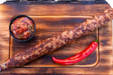 Uzun, mükemmel ızgara bir kebap ahşap bir tabakta duruyor, yanında da küçük bir kase baharatlı salsa ve canlı kırmızı biber var. Tabakta zengin lezzetler ve dokular sergileniyor.