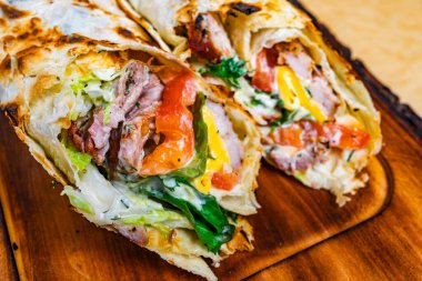 Izgara dürüm, taze et, taze sebze ve kremalı sos. Paket altın kahverengidir ve ahşap bir tahtanın üzerinde sunulur. İştah açıcı görünümünü vurgular..