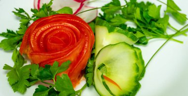 Taze kişniş ve salatalık dilimleriyle çevrili domatesten yapılmış bir gül içeren canlı bir garnitür gösterisi. Bu sanatsal sunum mutfağın yaratıcılığını ve tazeliğini gösteriyor.