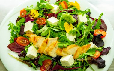 Karışık sebzeler, ızgara tavuk dilimleri, vişne domatesleri ve yenilebilir çiçeklerle süslenmiş beyaz bir tabakta servis edilen beyaz bir salata..