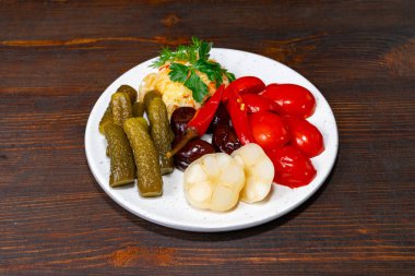 Beyaz bir tabağa yerleştirilmiş canlı bir sebze koleksiyonu. Etkinlikte kırmızı biber, domates, turşu ve taze otlarla süslenmiş sarımsak yer alıyor..