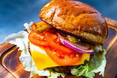 Tahtada servis edilen taze domates, marul, turşu, soğan ve peynirle kaplı lezzetli bir hamburger. Ekmek altın kahverengidir ve hafifçe parlaktır..