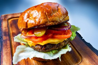 Lezzetli bir hamburgerde sulu bir köfte, erimiş peynir, ve taze sebzeler, marul, domates, soğan ve turşu gibi, ahşap bir kesme tahtasındaki parlak bir topuz üzerinde servis edilir..