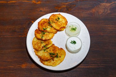 Altın rengi patatesli krepler, iki küçük kase kremalı sosun yanında düzgün bir şekilde servis edilir. Yeşil otlar yemeği süslüyor ve cazip bir sunum oluşturuyor..