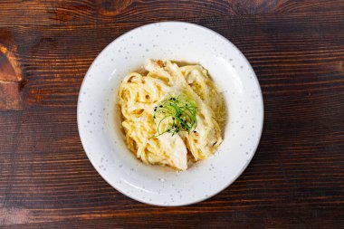 Taze otlarla süslenmiş taze tavuk parçaları içeren bir tabak kremalı makarna, ahşap bir masada oturur ve lezzetli bir yemek deneyimini davet eder..