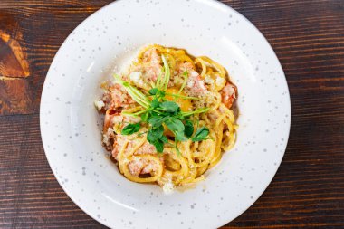 Taze otlarla kaplanmış bol kremalı makarna, benekli beyaz bir tabakta duruyor. Yemek zengin bir sos içeriyor ve zarif bir şekilde sunuluyor..