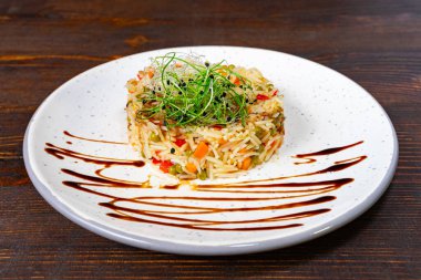 Üzerine koyu bir sos serpiştirilmiş renkli sebzeler ve otlarla güzel bir şekilde hazırlanmış pirinç servisi, beyaz bir tabakta ahşap bir arka planda zarif bir şekilde sunuluyor..