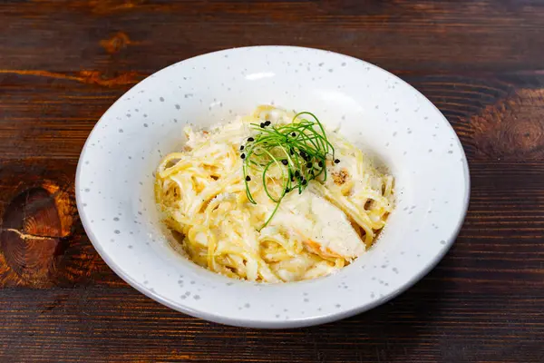 Süslü, beyaz bir kâse içinde taze otlarla süslenmiş kremalı bir makarna tabağı sunulur. Kırsal ahşap masa yemeğin davetkar atmosferini arttırır..