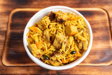 Kokulu pilav, taze tavuk parçaları ve canlı baharatlarla kokulu tavuk biryani ile dolu bir kase. Tahta bir yüzeye yerleştirilir, zengin renklerini ve dokularını ön plana çıkarır..