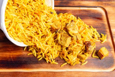 Canlı bir pirinç tabağı beyaz bir kaseden ahşap bir yüzeye dökülür, baharat ve yumuşak tavuk parçaları ile doldurulmuş kabarık taneler sergilenir. Bu iştah açıcı düzenleme doyurucu bir yemeği vurguluyor..