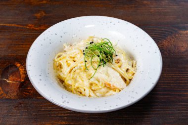 Süslü, beyaz bir kâse içinde taze otlarla süslenmiş kremalı bir makarna tabağı sunulur. Kırsal ahşap masa yemeğin davetkar atmosferini arttırır..