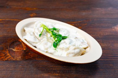 Erimiş hamur köfteleri, taze otlarla süslenmiş kremalı bir sosun içine yerleştirilir, kırsal bir ahşap masada oval bir tabakta servis edilir, sıcak ve davetkar bir yemek deneyimi yaratır..