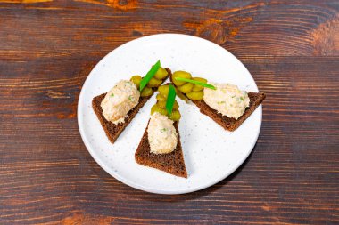 Kremalı ve turşuyla süslenmiş küçük üçgen çavdar ekmeği parçaları, sofistike bir aperatifin altını çizerek beyaz bir tabakta güzelce yerleştirilmiştir..