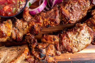 Sulu ızgara et parçaları, taze kırmızı soğanlar ve bir kase canlı salsa eşliğinde ahşap bir tabakta düzenlenir. Bu yemek her barbekü için mükemmel lezzetler sunuyor..