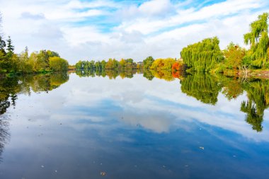 A peaceful river landscape showcases vibrant autumn colors along the banks. The clear water reflects the trees and clouds, creating a serene atmosphere on a calm day.
