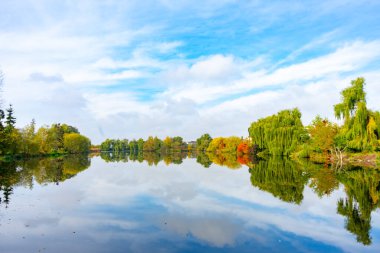 Parlak sonbahar renkleri, berrak bir gökyüzünün altındaki canlı ağaçları yansıtan huzurlu bir gölü sarıyor. Bu dingin ortam, sonbahar boyunca doğanın güzelliğinin rahatlaması ve takdir edilmesini sağlıyor..