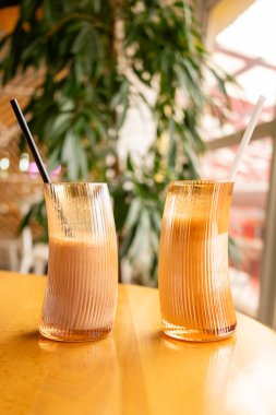 Canlı bir kafede iki renkli içecek bir masanın üzerine konur. Bir içecek kremalı ve kahverengidir, diğeri ise açık ve turuncu. Parlak bitkiler arka planı çerçeveletip, rahat atmosferi güçlendirir.