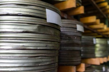 Metalik film makaraları loş aydınlatılmış bir depodaki ahşap raflarda duruyor. Makaraların karmaşık detayları geçmişten gelen hikayelerin keşfedilmeyi beklediğini gösteriyor..