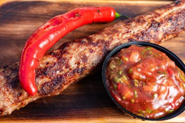 Tahta bir tahtanın üzerinde nefis bir kebap duruyor, yanında küçük bir kâse içinde baharatlı bir sos ve canlı kırmızı biberle süslenmiş, et severler için mükemmel bir yemek sergiliyor..