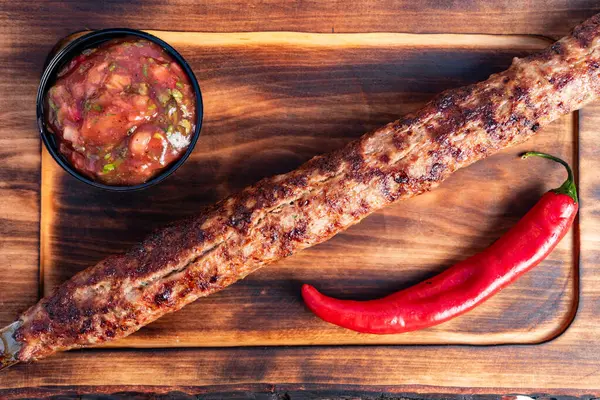 Uzun ızgara bir kebap, bir kase baharatlı salsa ve canlı kırmızı biberin yanında ahşap bir tabakta duruyor. Lezzetli ve iştah açıcı bir yemek sunuyor..