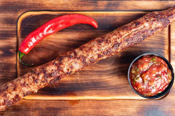 Sulu kebap ahşap bir tahtanın üzerinde duruyor, yanında da bir kase baharatlı salsa ve canlı kırmızı biber var. Günün her saatinde lezzetli bir yemek için mükemmel..