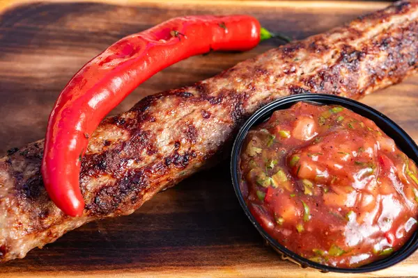 Tahta bir tahtanın üzerinde nefis bir kebap duruyor, yanında küçük bir kâse içinde baharatlı bir sos ve canlı kırmızı biberle süslenmiş, et severler için mükemmel bir yemek sergiliyor..
