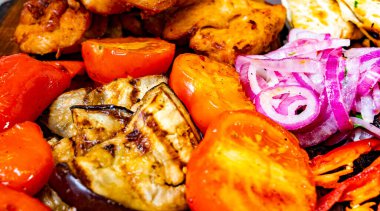 Canlı bir tabakta ızgara domates, patlıcan, soğan ve baharatlı etler yer alıyor. Tabakta taze renkler ve paylaşımı ve zevki davet eden ağız sulandırıcı bir sunum sergileniyor.