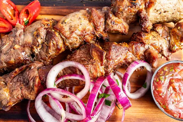 Lezzetli ızgara et şişleri, canlı kırmızı soğanlar, taze acı biberler ve biraz da baharatlı sos ile servis edilir. Tahtadaki sunum yemeğin iştah açıcı görünümünü arttırır..