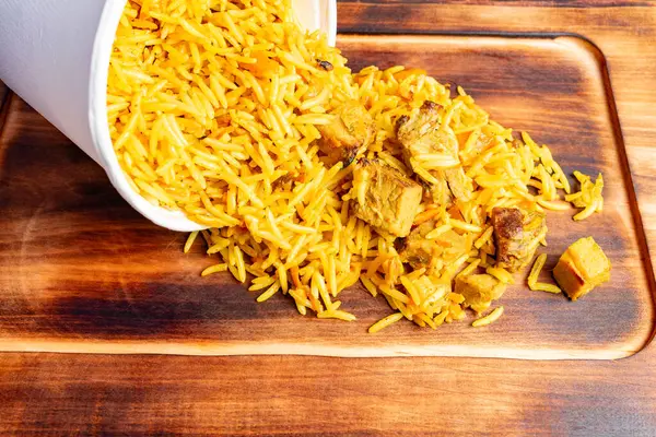 Yumuşak etle karıştırılmış sarı pirinç beyaz bir konteynırdan ahşap bir yüzeye dökülüyor. Canlı renk ve doku davetkar bir mutfak görüntüsü yaratır..