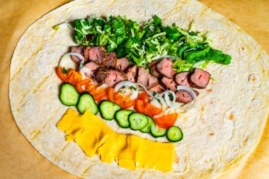 Bir mutfak hazırlığı dilimlenmiş et, marul, domates, soğan, salatalık ve peynirle dolu bir tortilla gösteriyor. Canlı renkler iştah açıcı bir görsel oluşturur..