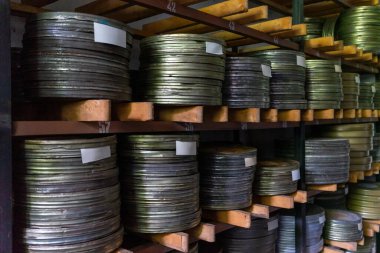Metalik film yığınları, loş bir film arşivindeki ahşap raflarda düzgün bir şekilde dizilmiş. Filmdeki etiketler içeriklerini gösteriyor, nostaljik bir atmosfer yaratıyor..