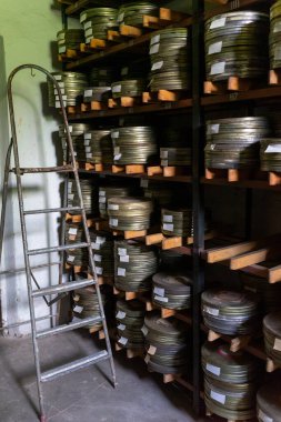 Arşiv odasındaki ahşap raflarda düzgünce dizilmiş sayısız film makarası var. Rafların yanında duran metal bir merdiven daha üst düzey bir depoya erişimi gösteriyor..