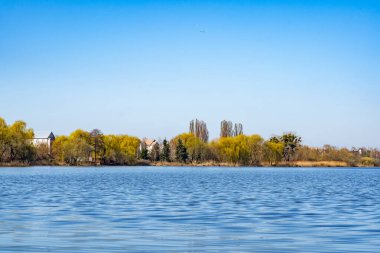 Sakin bir göl, bereketli ağaçların kıyılarına çizdiği berrak mavi gökyüzünü yansıtır. Yakınlardaki evler yeşillikleri gözetliyor. Bu da sonbahar boyunca huzurlu ve kırsal bir ortam olduğunu gösteriyor..