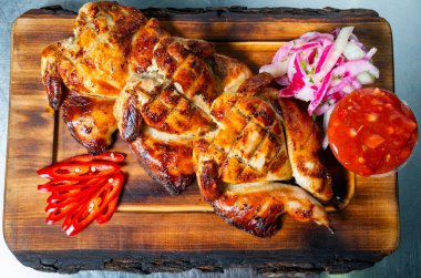 Lezzetli ızgara tavuk parçaları ahşap bir tahtanın üzerinde kaplıdır. Yanında dilimlenmiş soğan, baharatlı kırmızı biber ve küçük bir kase keskin sos ile lezzetli bir ziyafet..