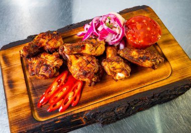 Izgara tavuk kanatları, dilimlenmiş kırmızı soğanlar, taze otlar ve baharatlı sosla süslenmiş ahşap bir tabakta düzenlenir. Canlı kırmızı biberler yemeğe renk katar..