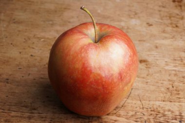 Kırsal bir ahşap tezgahın üzerinde yalnız kırmızı bir elma oturuyor. Yumuşak gün ışığında pürüzsüz cildi, elmanın canlı renklerini ve davetkar görünümünü aydınlatır..