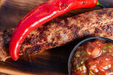 Lezzetli ızgara sosis yanında canlı kırmızı biber ve küçük bir kase taze salsa sosu servis edilir. Düzenleme sıcak ahşap bir tahta üzerine yerleştiriliyor, kırsal hissi arttırıyor..