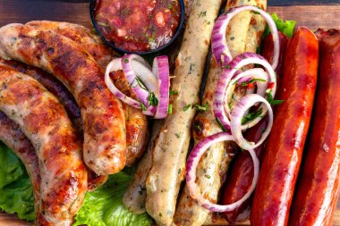 Ahşap bir tahtanın üzerinde canlı bir ızgara sosis karışımı. Tabakta taze salsa sosu ve ince dilimlenmiş soğanlarla birlikte çeşitli tatlar yer alıyor ve bu da iştah açıcı bir sunum oluşturuyor..