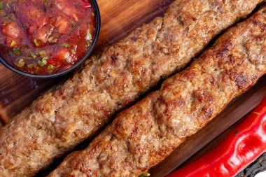 İki ızgara kebap ahşap bir tahtanın üzerinde, güzelce kaşarlanmış ve hafifçe yanmış. Onlara domates ve otlarla dolu küçük bir kase enerjik salsa sosu eşlik eder..