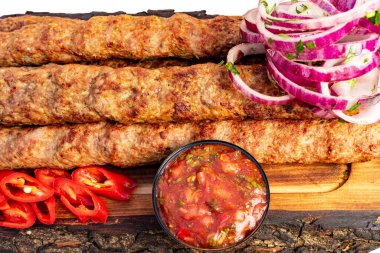 Izgara kebaplar canlı kırmızı soğanlar, taze biber ve bir kase baharatlı salsa sosuyla birlikte ahşap bir tahtanın üzerinde sunulur. Bu yemek zengin tat ve renkleri vurgular.