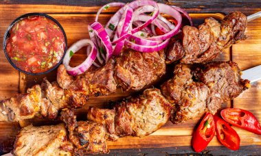 Taze salsa, dilimlenmiş soğan ve kırmızı biberle birlikte ahşap bir tepside duran tuzlu ızgara et şişleri doyurucu ve iştah açıcı bir yemek oluşturuyor..