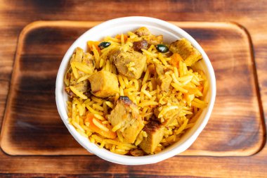 Sıcak bir kase sarı pirinç, yumuşak tavuk parçaları, renkli sebzeler ve aromatik baharatlarla karıştırılmış ahşap bir yüzeye oturmuş, doyurucu bir yemeği davet ediyor..