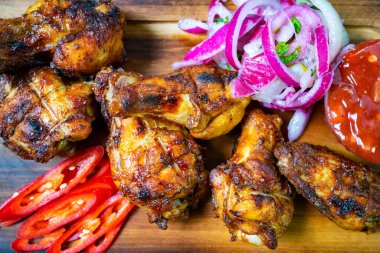 Izgara tavuk kanatları ahşap bir tabakta, kırmızı biber dilimleri, keskin soğanlar ve yanında domates sosu ile güzelce düzenlenerek iştah açıcı bir gösteri sunuluyor..