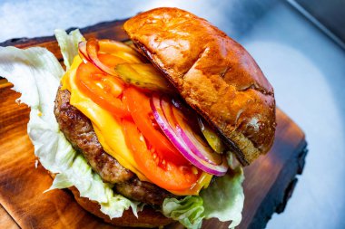 Ağız sulandıran bir hamburger, içinde bir biftek köftesi, çedar peyniri, marul, domates, turşu ve soğan var. Tahta bir tahtanın üzerinde. Canlı renkler ve katmanlar lezzetli bir ısırık davet ediyor..