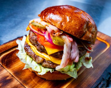 Eritilmiş peynir, pastırma, taze marul, domates, turşu ve soğanla dolu sulu bir hamburger, ahşap servis tahtasının üzerinde duruyor..