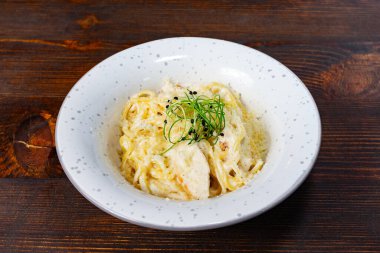 Kremalı makarna tabağı zarif bir şekilde taze otlarla süslenmiş beyaz bir kaseyle sunulur. Ahşap masa kırsal bir cazibe katıyor. Sıcak bir yemek deneyimi için mükemmel..