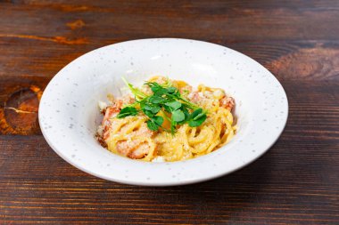 Taze ot ve peynirle kaplanmış bir spagetti kasesi ahşap bir masada duruyor. Yemek davetkar ve zevk almaya hazır. Kremalı sosunu ve cömert porsiyonlarını sergiliyor..