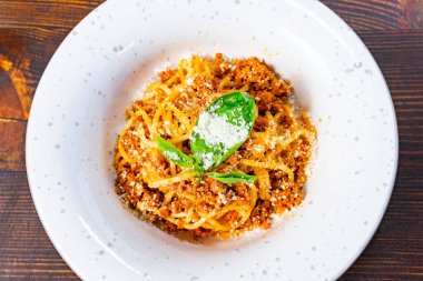 Zengin et soslu bir tabak spagetti, rendelenmiş peynir ve ahşap bir masada taze bir fesleğen yaprağı duruyor. Yemek canlı renkler ve iştah açıcı dokular sergiliyor..